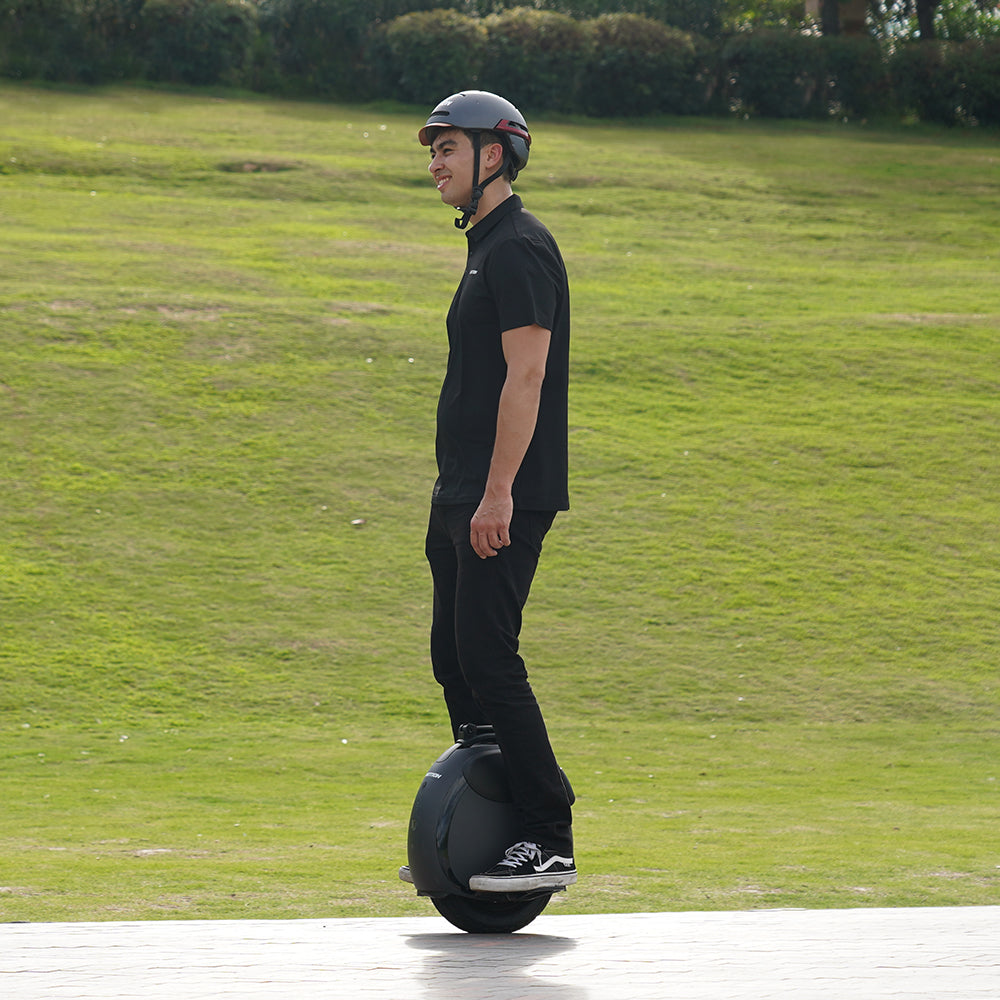 A man rides the INMOTION V6 electric unicycle on a paved path, wearing a helmet and black outfit.