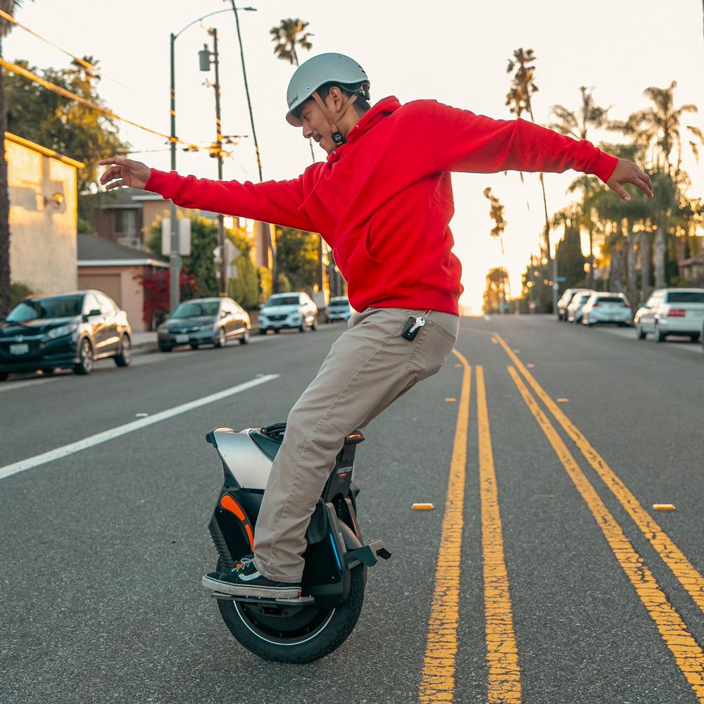 A man in a red hoodie rides a V12S electric unicycle on a sunny street, balancing with arms outstretched.
