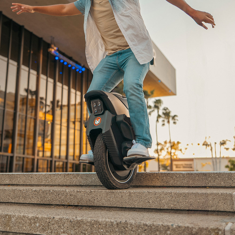 A person rides a V12S electric unicycle, balancing on a curb with arms outstretched in an urban setting.